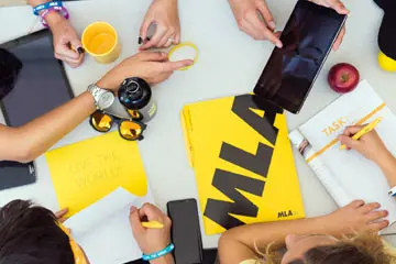 Overhead view of students at a table with MLA branded notebooks, phones and coffee