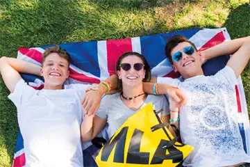 Three teenagers lying on a Union Jack flag on grass with an MLA towel, smiling