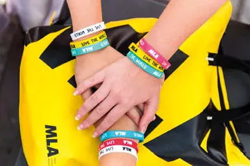 Stacked hands wearing colourful MLA "Live the MLA" wristbands on a yellow bag
