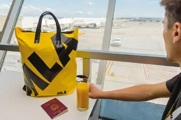 Boy at an airport gate with a yellow MLA bag, passport and drink, plane outside