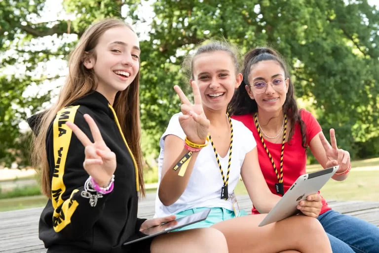 Three smiling girls with MLA lanyards making peace signs while holding tablets in a park