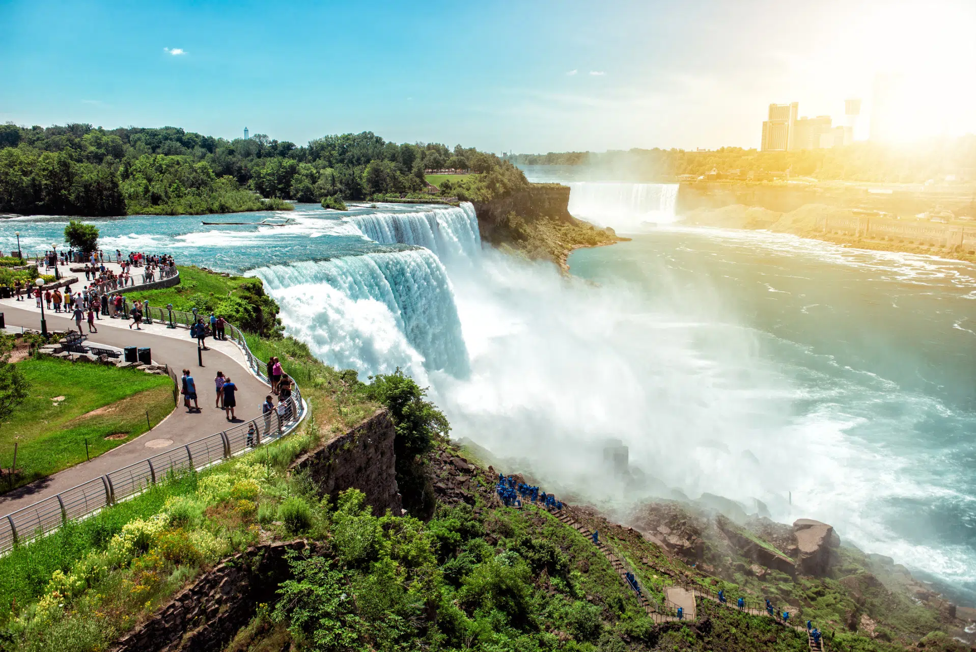 Niagara Falls with visitors on a viewing walkway, rushing water and mist under a sunny sky