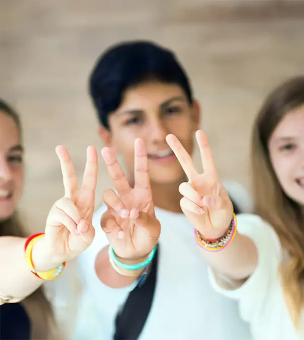 Three teenagers smiling and holding up victory hand signs with colourful friendship bracelets